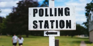 A weathered white sign with the words "POLLING STATION" and an arrow pointing left, in front of a blurred background featuring two people walking in a park.