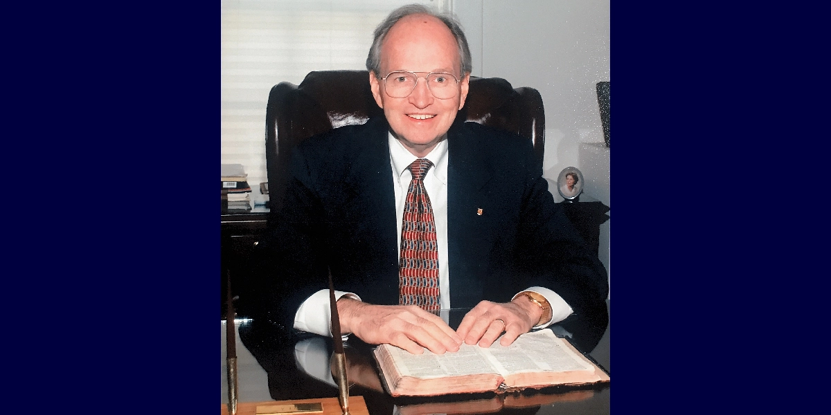 Pastor Robert L. Alderman seated at a desk with an open Bible and pens.